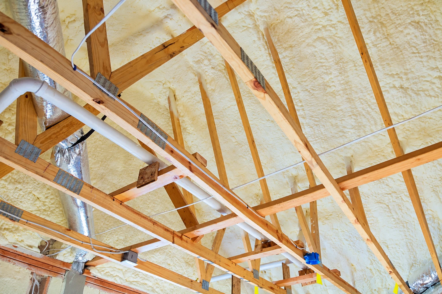Yellow spray foam fills the roof cavities among exposed wooden trusses, pipes and silver ductwork in an attic.