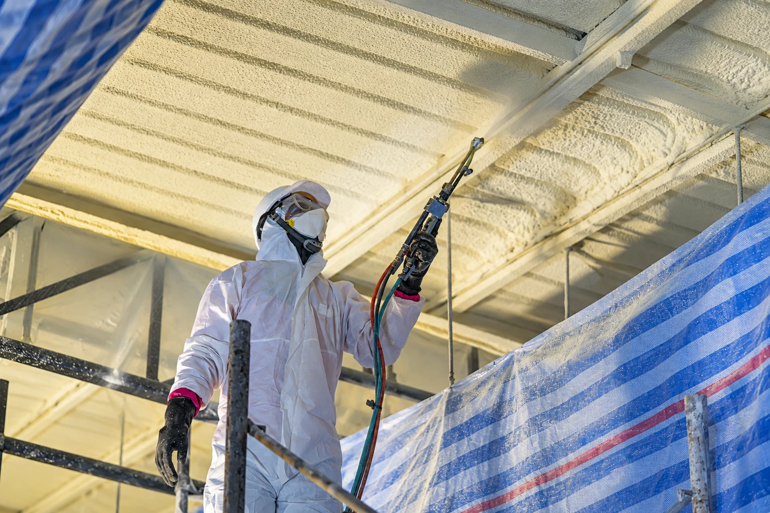 Worker in protective gear applies foam insulation to a ceiling with a spray gun.
