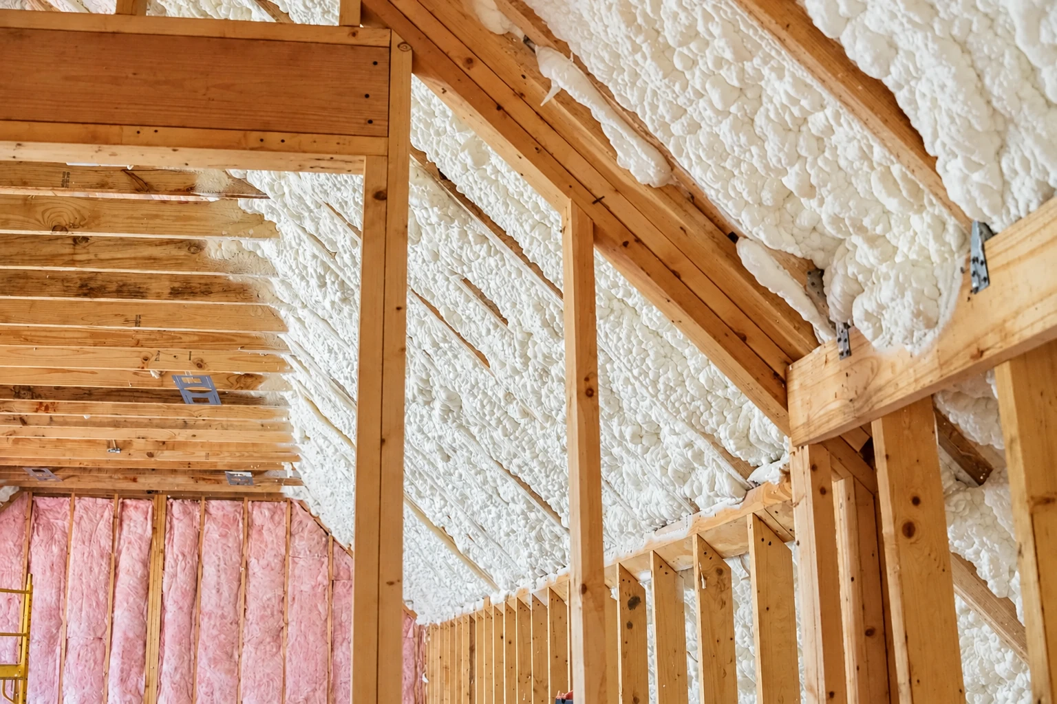 White spray foam covers the sloped roof between exposed wooden rafters in an unfinished building.